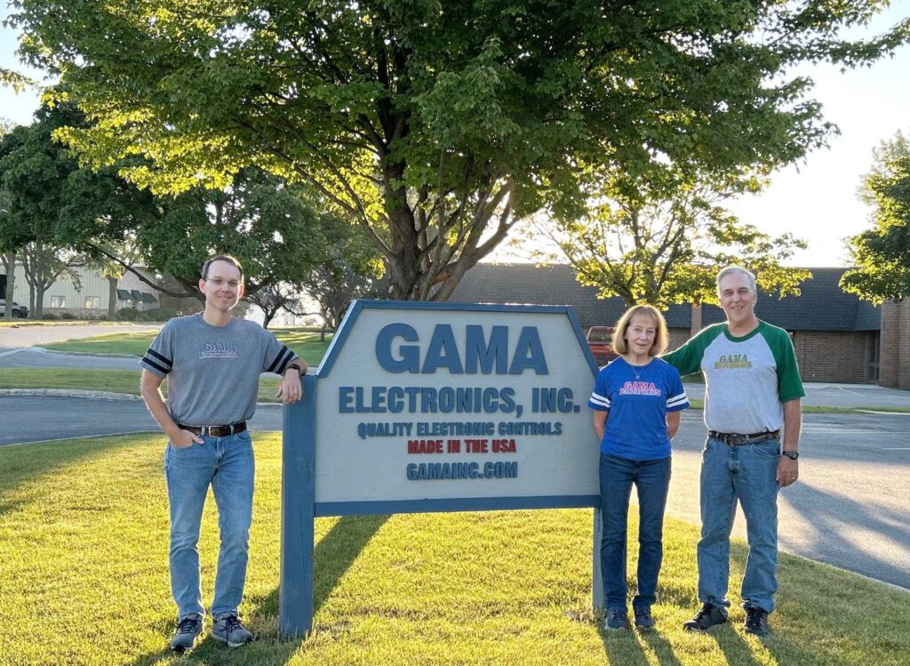 Leadership team members standing around a sign for GAMA Electronics, Inc., outside on a grassy area with trees and a building in the background.